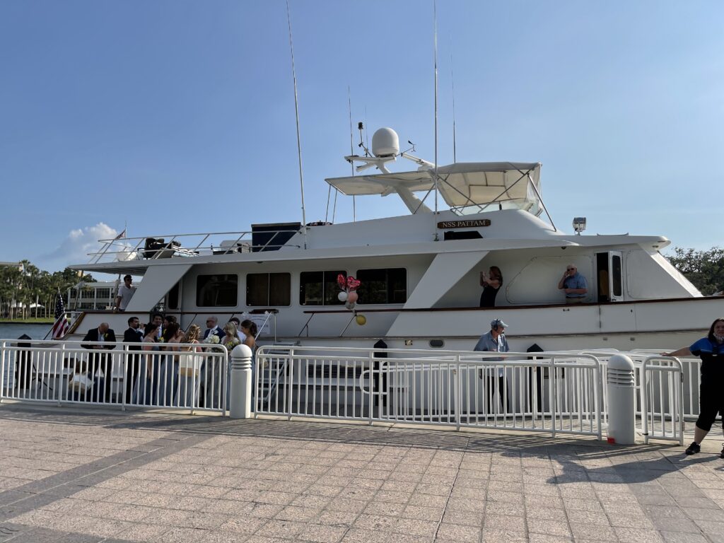 A group of passengers boarding a white yacht named "Sensation" docked at a pier with railings on a sunny day for a dolphin cruise.