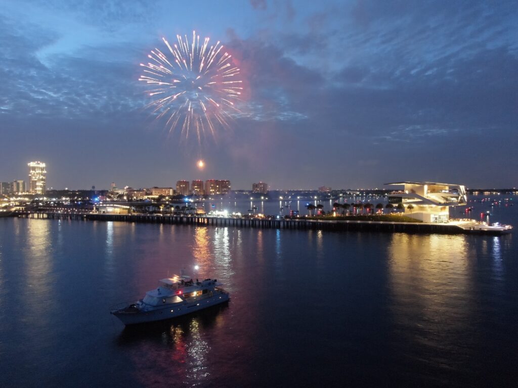 Aerial view of a yacht on the water near a city waterfront, with fireworks lighting up the evening sky in Tampa.