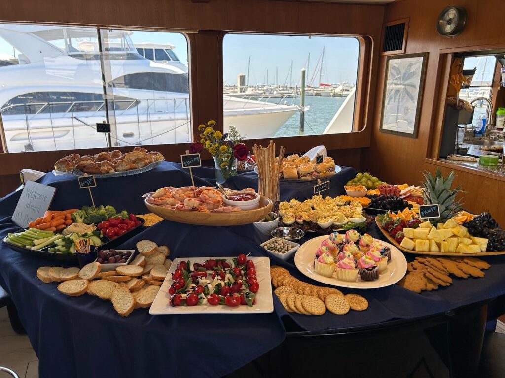 A spread of assorted appetizers and desserts on a table inside a yacht with a view of the marina through the windows.